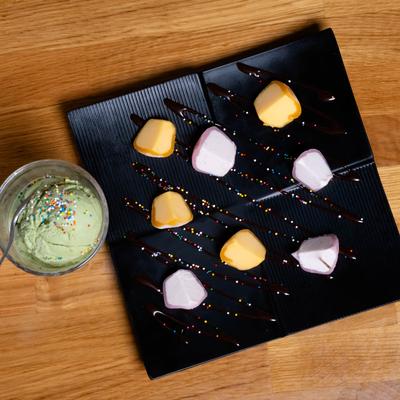 Square black plate with yellow and pink mochi next to a glass of green tea ice cream.