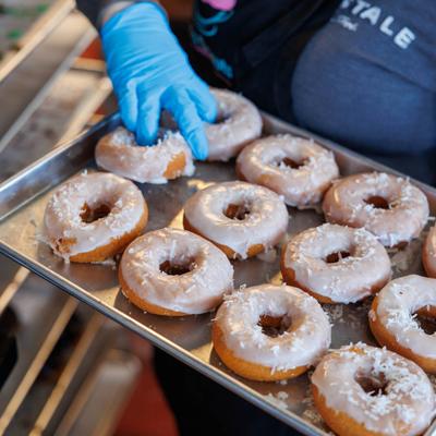 A hand in a blue glove holding a tray of glazed donuts topped with shredded coconut.