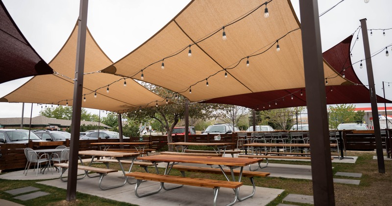 Patio area, long tables with bench seating and chairs, covered with shade sails, lanterns