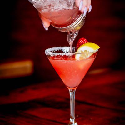 A pink cocktail in a sugar-rimmed margarita glass, garnished with a lemon slice and strawberry.