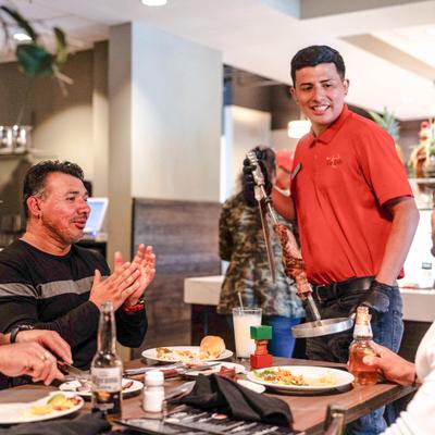 A server is presenting grilled meats on a large skewer to diners.