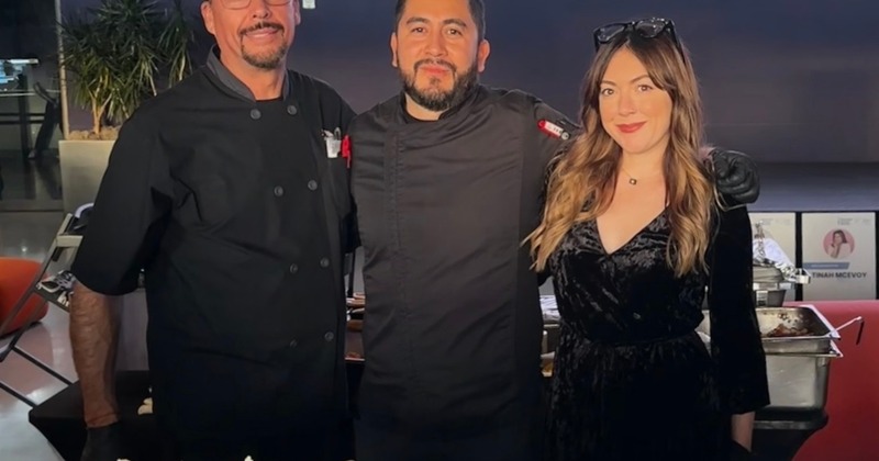 Three staff members pose in indoor space with catering trays behind them