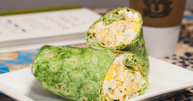 A green spinach wrap cut in half, filled with egg salad, sits on a white plate