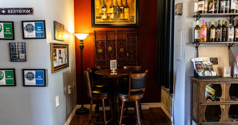 A cozy interior corner with a table and chairs, framed awards, a floor lamp, wine bottles