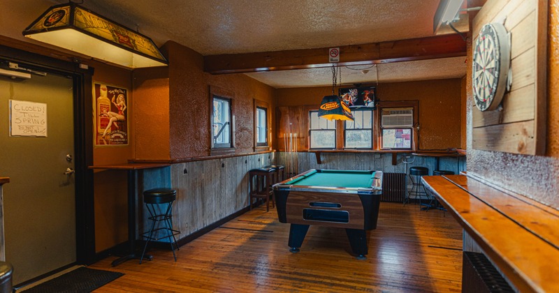 Interior with wooden flor and walls, pool table, dartboard and stained glass billiard light fixtures