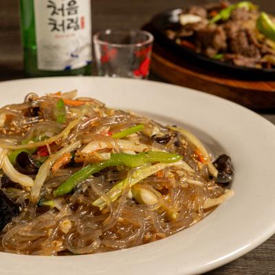 Plate of stir-fry glass noodles, alongside bottle of sake and other food items.