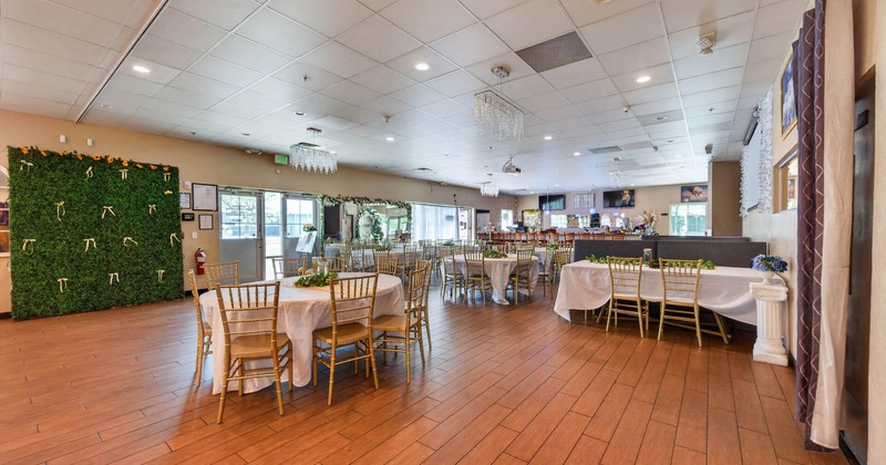 A large banquet hall with round tables and golden chairs, wooden flooring, and decorative greenery