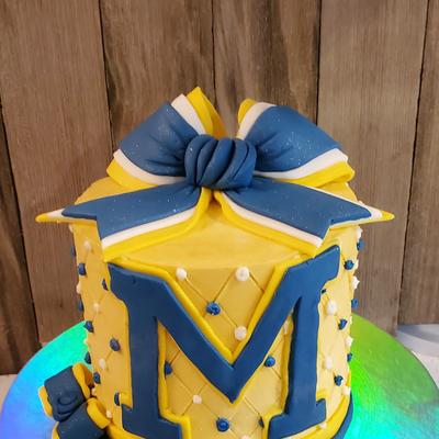 A round cake with a  blue and yellow bow on top and a prominent blue letter M on the side.