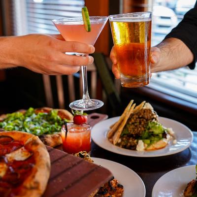 Interior, two people clink beer and cosmopolitan over a table with assorted dishes.