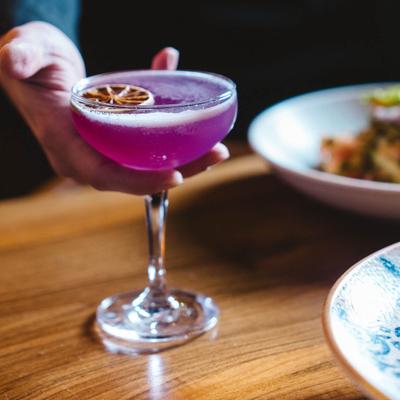 A hand holding a vibrant purple cocktail, set on a table with food plates.