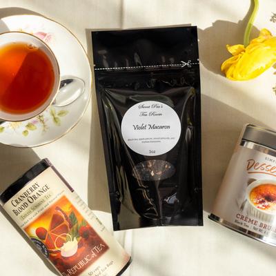 Black pouch of Violet Macaron tea surrounded by tea tins and a teacup.