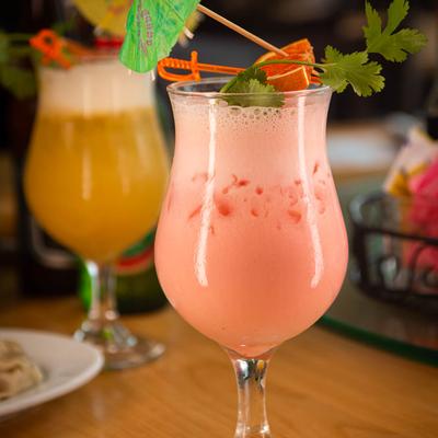 A pink cocktail with ice in a glass, topped with garnish, sits on a wooden table.