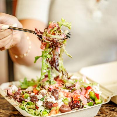 A person lifting a forkful of Greek salad from a takeout container.