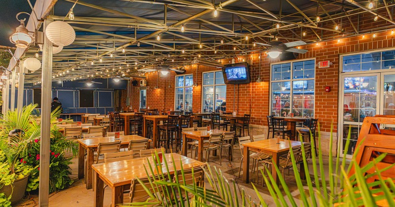 Outdoor restaurant patio with wooden tables, string lights, and a brick wall with windows and a TV