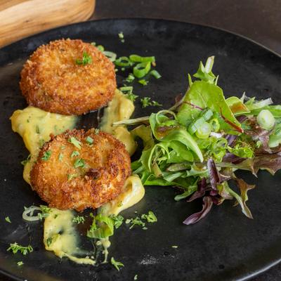 Two golden-brown crab cakes sit on a dark plate with sauce, accompanied with a green salad.