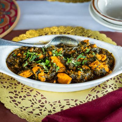 Chicken Saag in a white oval dish atop a golden doily on a red table.