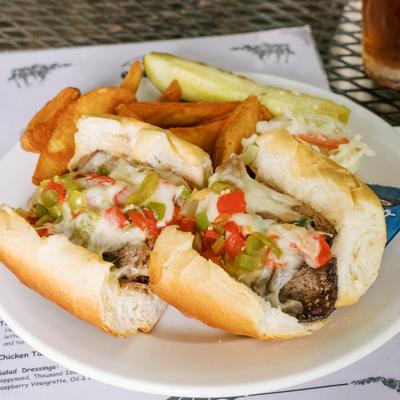 A Philly cheesesteak sandwich served with French fries and a crisp pickle spear.