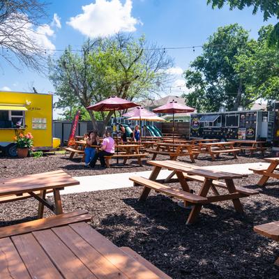 Outdoor seating area with picnic tables, food trucks, and umbrellas on a sunny day.