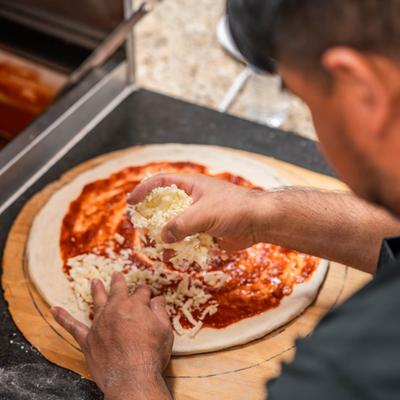 Pizza chef adding shredded mozzarella to a pizza base topped with tomato sauce.