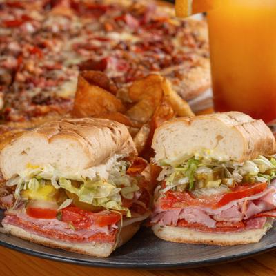 Italian sub sandwich served with chips, with a pizza and a drink in the background.