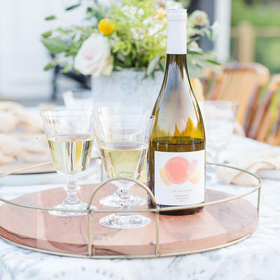 A bottle of white wine and two glasses sit on a wooden tray outdoors