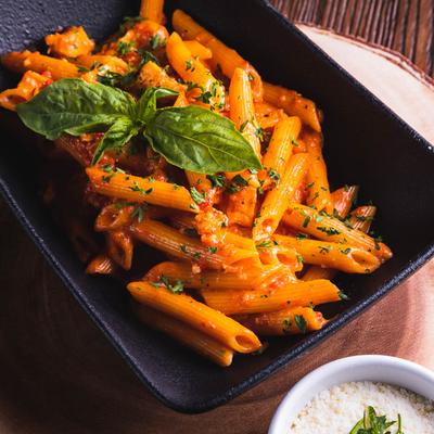 A black dish filled with penne pasta in a rich tomato sauce, garnished with fresh basil and parsley.