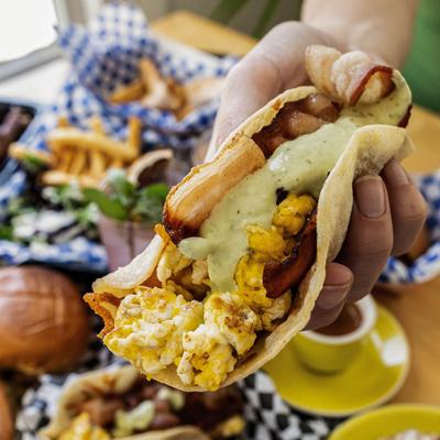 Breakfast taco in a hand, with assorted food plates in blurred background.