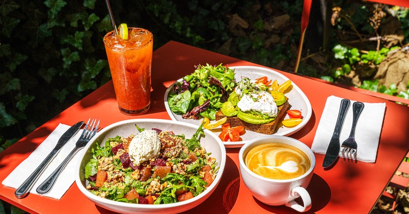A quinoa salad, avocado toast with greens, a latte, and a Bloody Mary displayed outdoors