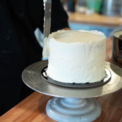 Employee uses a spatula to smoothly frost a round cake with white icing.