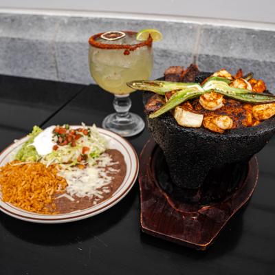 Molcajete dish served in volcanic stone bowl beside separate plate of sides and a Margarita.