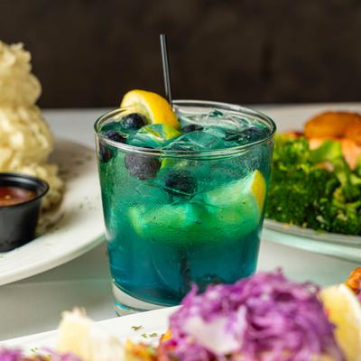 Blue cocktail with blueberries and lemon slices, surrounded by colorful dishes.