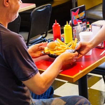 A guest sits at a red table in a diner receiving a tray of crinkle-cut fries and a burger.