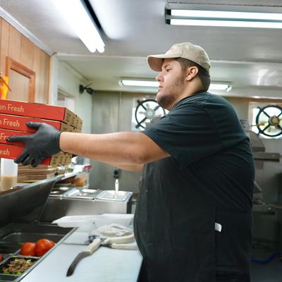 A worker stacking pizza boxes in a busy kitchen.