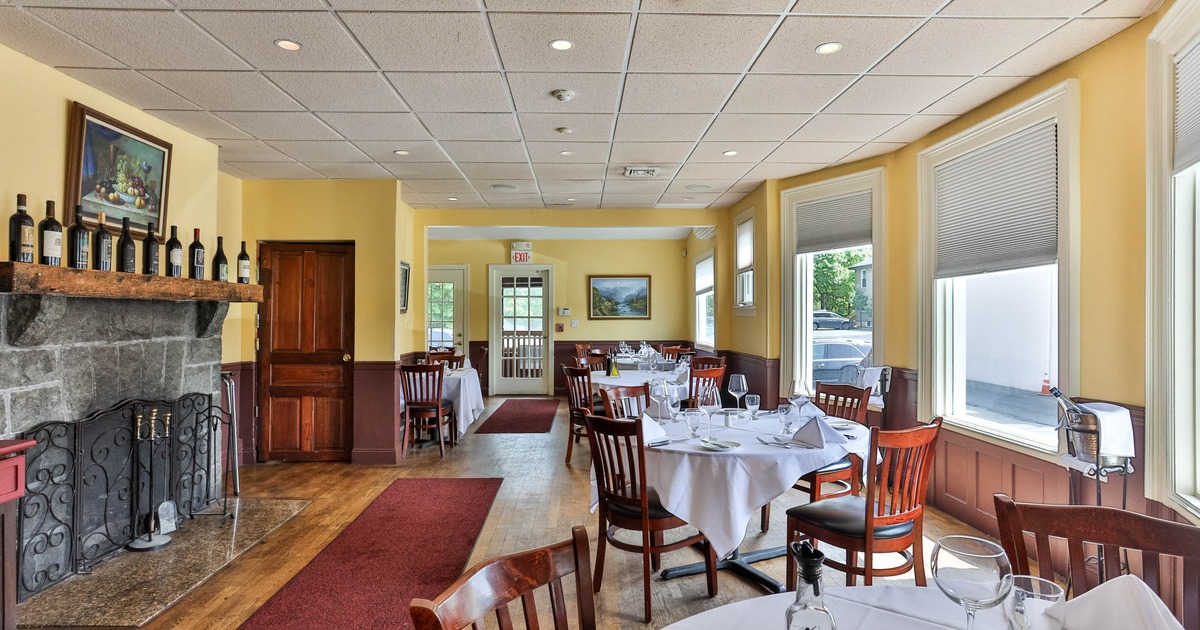 Interior of a restaurant with round tables, wooden chairs, stone fireplace, and large windows