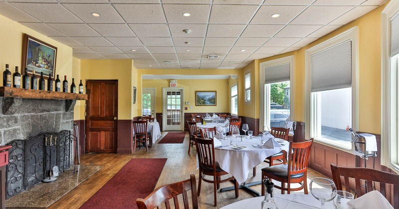 Interior of a restaurant with round tables, wooden chairs, stone fireplace, and large windows