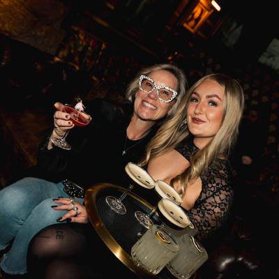Two smiling people pose in a dimly lit bar with a tray of cocktails.