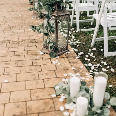 A rustic lantern with greenery on a pathway with white petals and candles.
