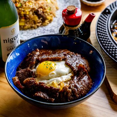 Blue bowl of rice topped with glazed beef, a fried egg and chopped green onions..