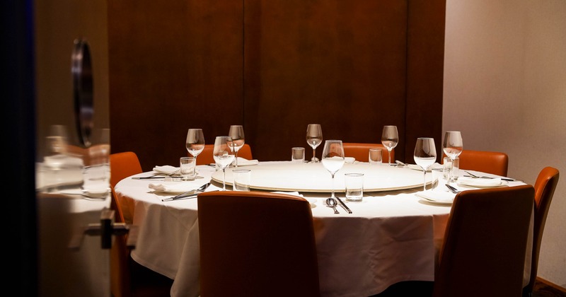 A round table set with wine glasses and cutlery, surrounded by brown chairs in a warmly lit room