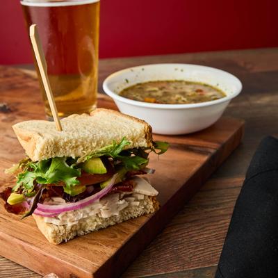 Turkey sandwich with lettuce, avocado, and red onion, served with a soup and a beer.