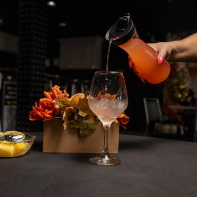Person pouring a orange beverage from a pitcher into a wine glass filled with ice.