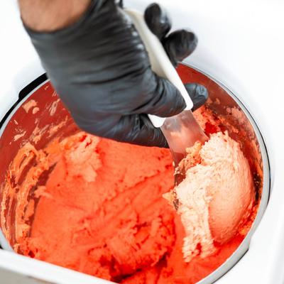 A hand in black gloves scooping  red gelato from a large metal container.