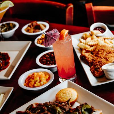 Tall orange cocktail with citrus and paper umbrella garnish surrounded by plated dishes and sides.