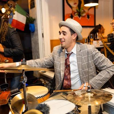 Musician wearing a plaid blazer and hat playing drums during live performance.