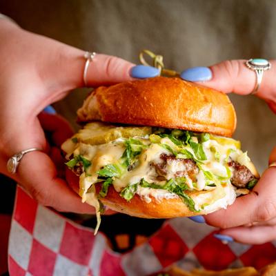 Hands holding a cheeseburger with lettuce, pickles, and sauce.