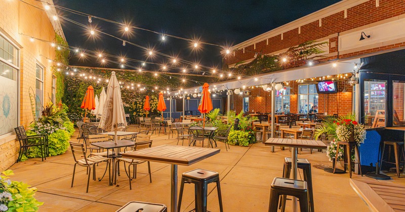 Outdoor patio at night with string lights, tables, chairs, and orange umbrellas