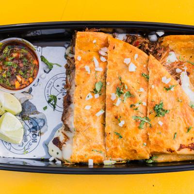Quesabirria served in a black take-out container with sides of consome and lime wedges.
