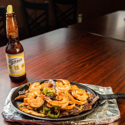 Shrimp and steak fajitas served sizzling on a cast-iron skillet alongside a bottle of beer.