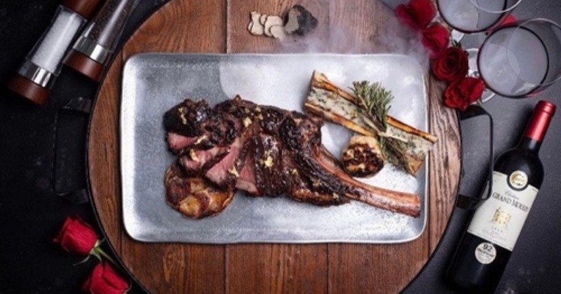 A plate with Tomahawk steak and bone marrow displayed with glasses, roses and a wine bottle