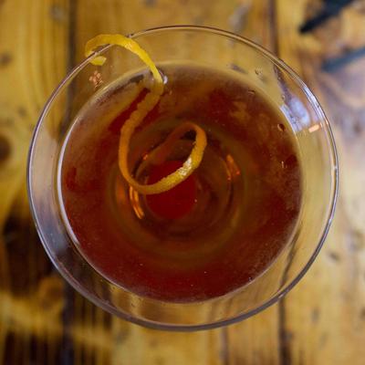 Amber cocktail with citrus twist and cherry centered in a glass on a wooden table.